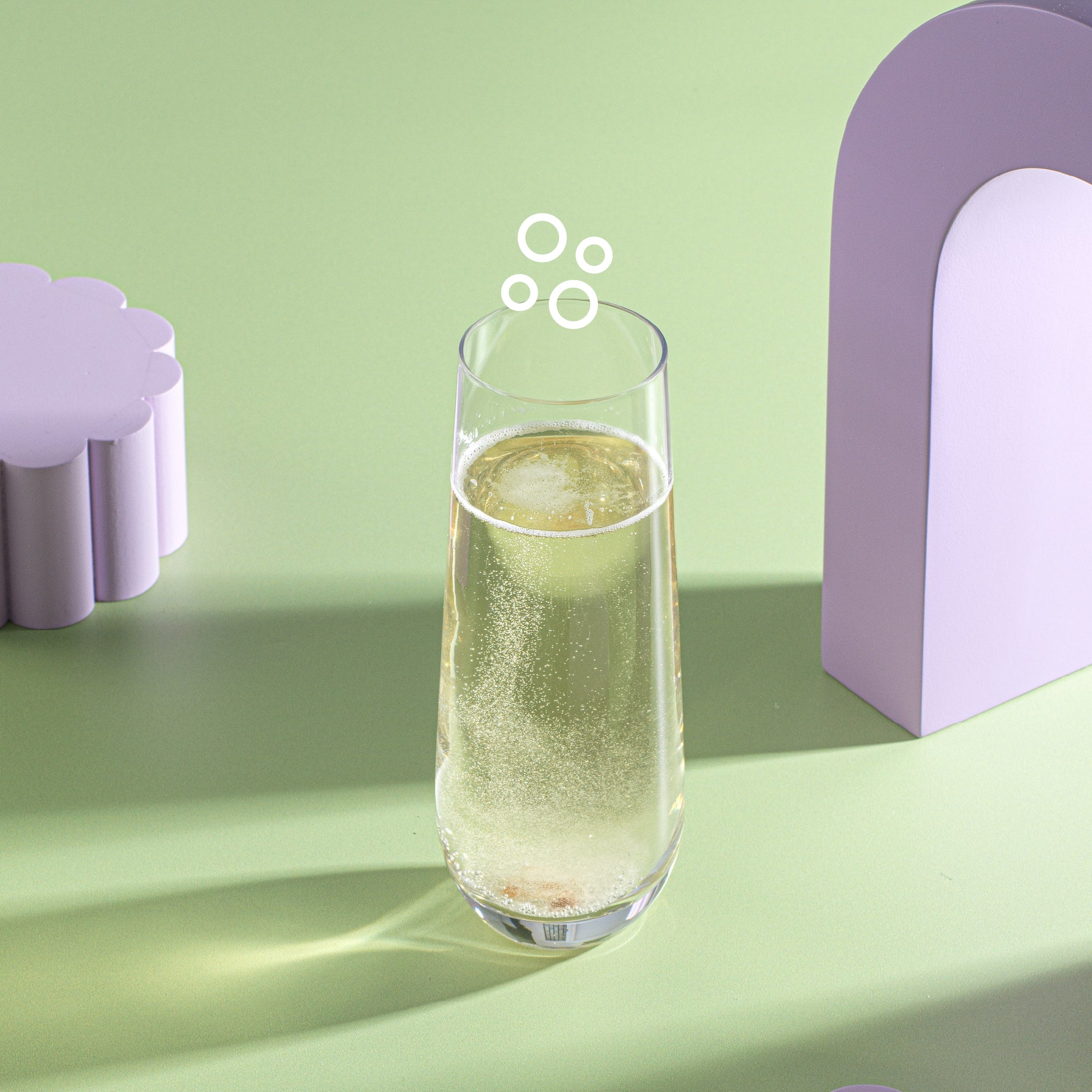 A Milo Champagne Glass on a green surface. There are two abstract shapes, both purple in the background.