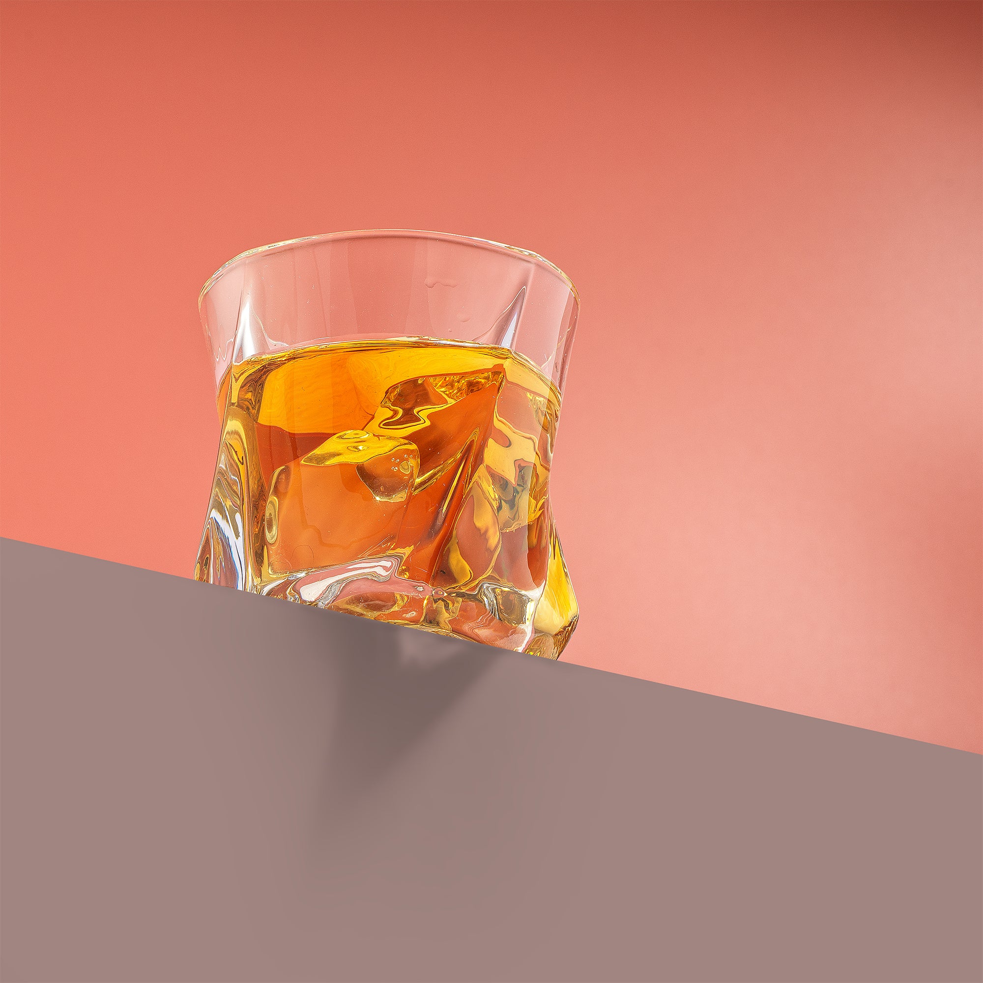 A twisted double old fashion glass filled with whiskey and ice on a ledge with a red background.
