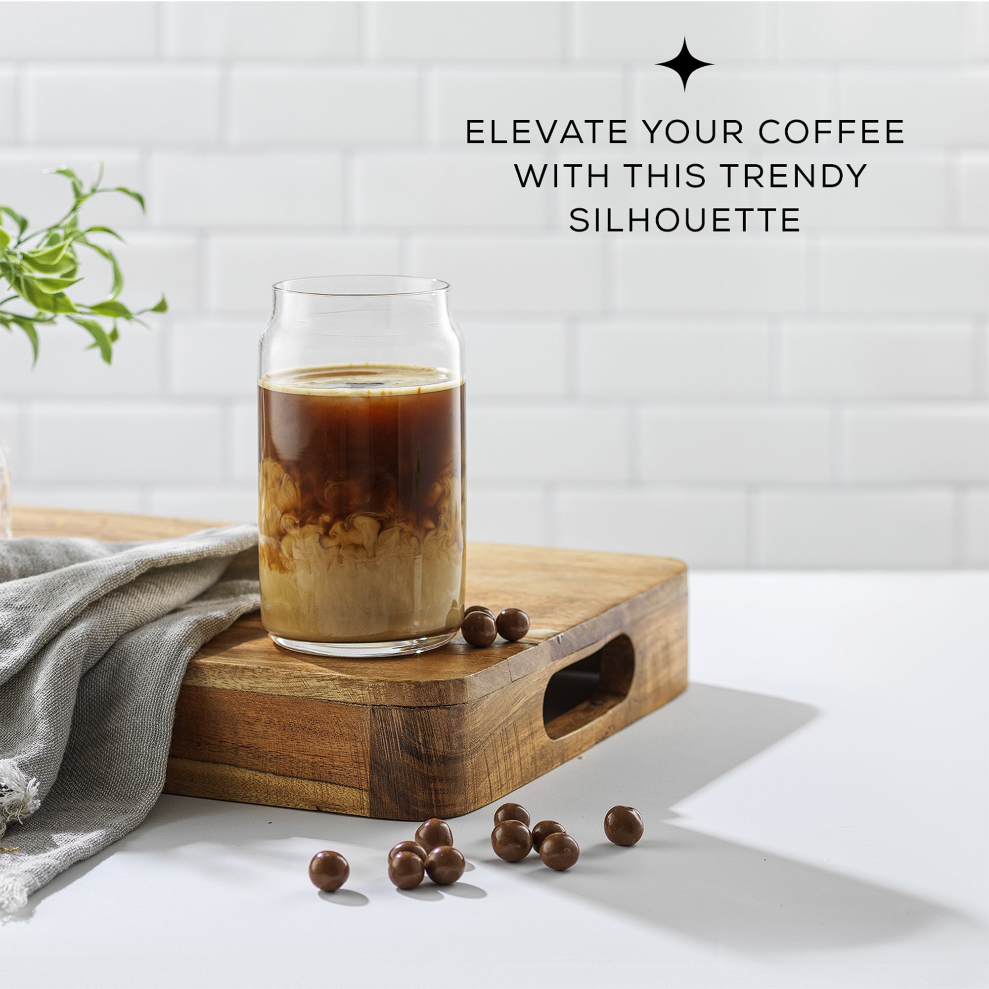 A JoyJolt glass of iced coffee with cream swirling on a wooden tray, surrounded by coffee beans, against a tiled background.