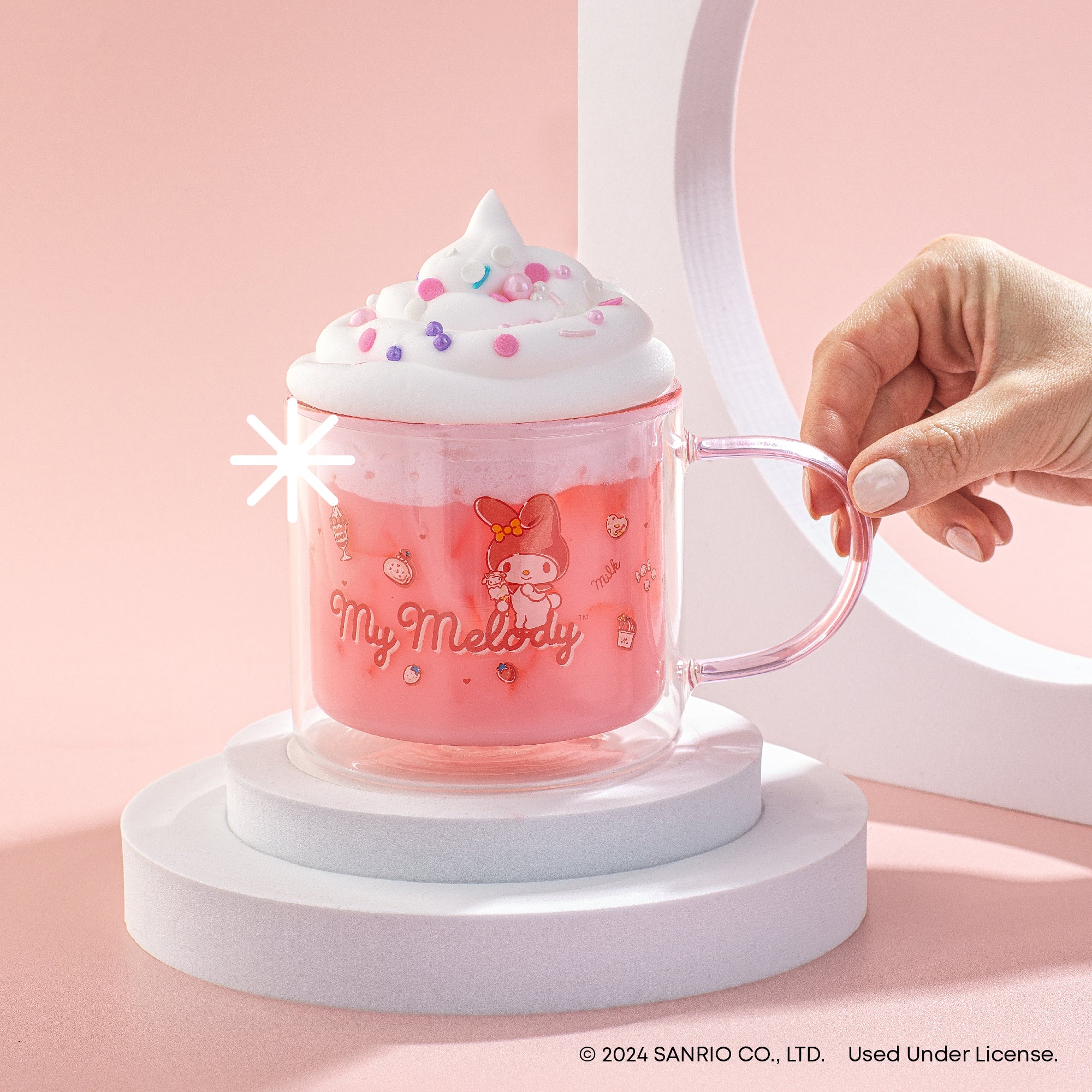A person holding a pink mug with whipped cream on top. The mug depicts My Melody. Text on the mug reads "My Melody" and includes Sanrio copyright information.