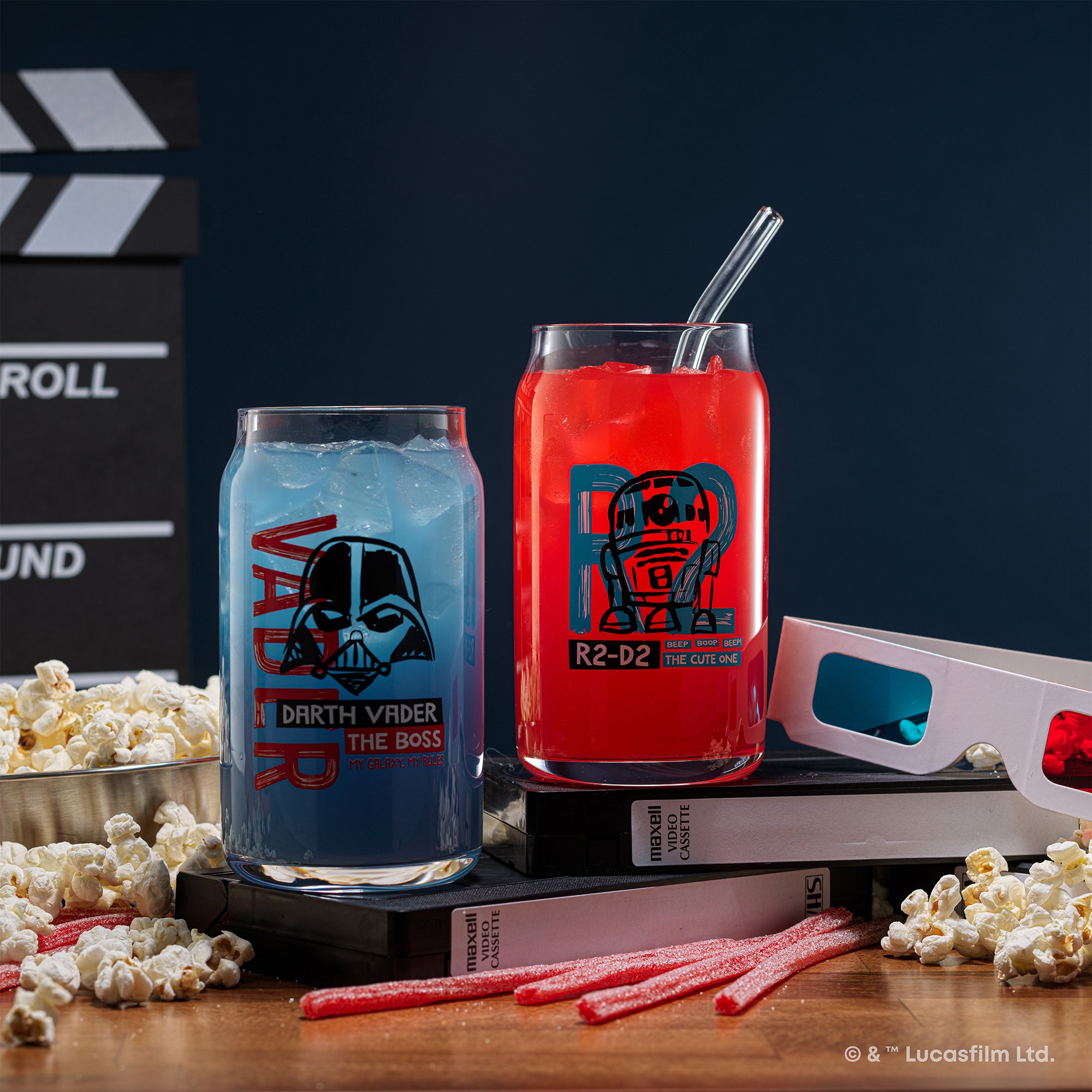 JoyJolt's Star Wars 'Now Playing' glass tumbler set featuring Darth Vadar and R2-D2. R2-D2 glass is on two VHS tapes and 3D glasses next to it. The glasses are surrounded by popcorn and sour straw candy.