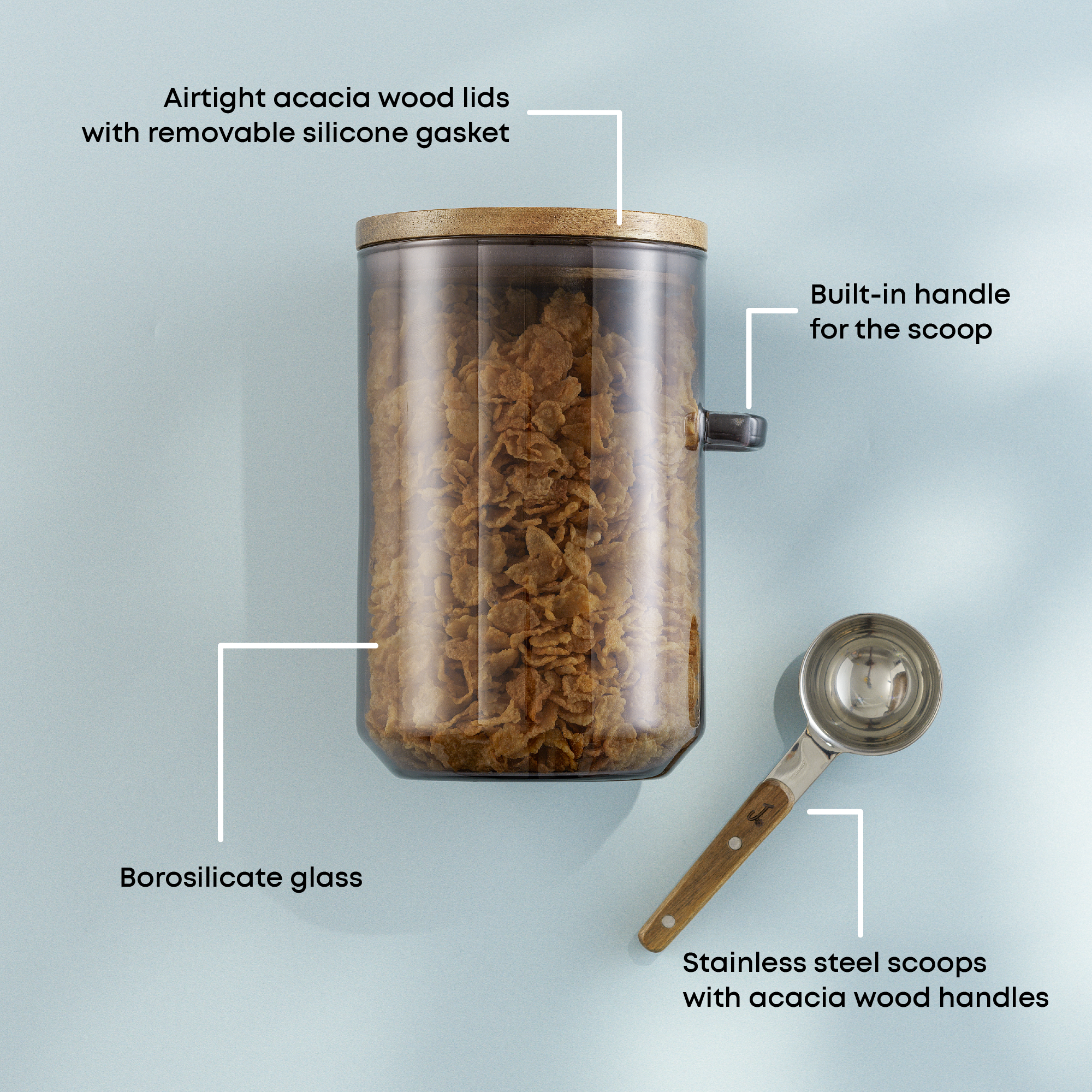 Glass jar with cereal, a measuring spoon, and text labels on a light blue background: airtight acacia lids, build in handle for scoop, borosilicate glass, scoops have wood handles and stainless steel scoops.