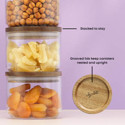 Three glass canisters with wooden lids, each containing different food items, on a light purple background. With text "stacked to stay, grooved lids keep canisters nested and upright"