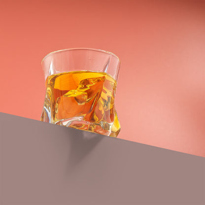 A twisted double old fashion glass filled with whiskey and ice on a ledge with a red background.