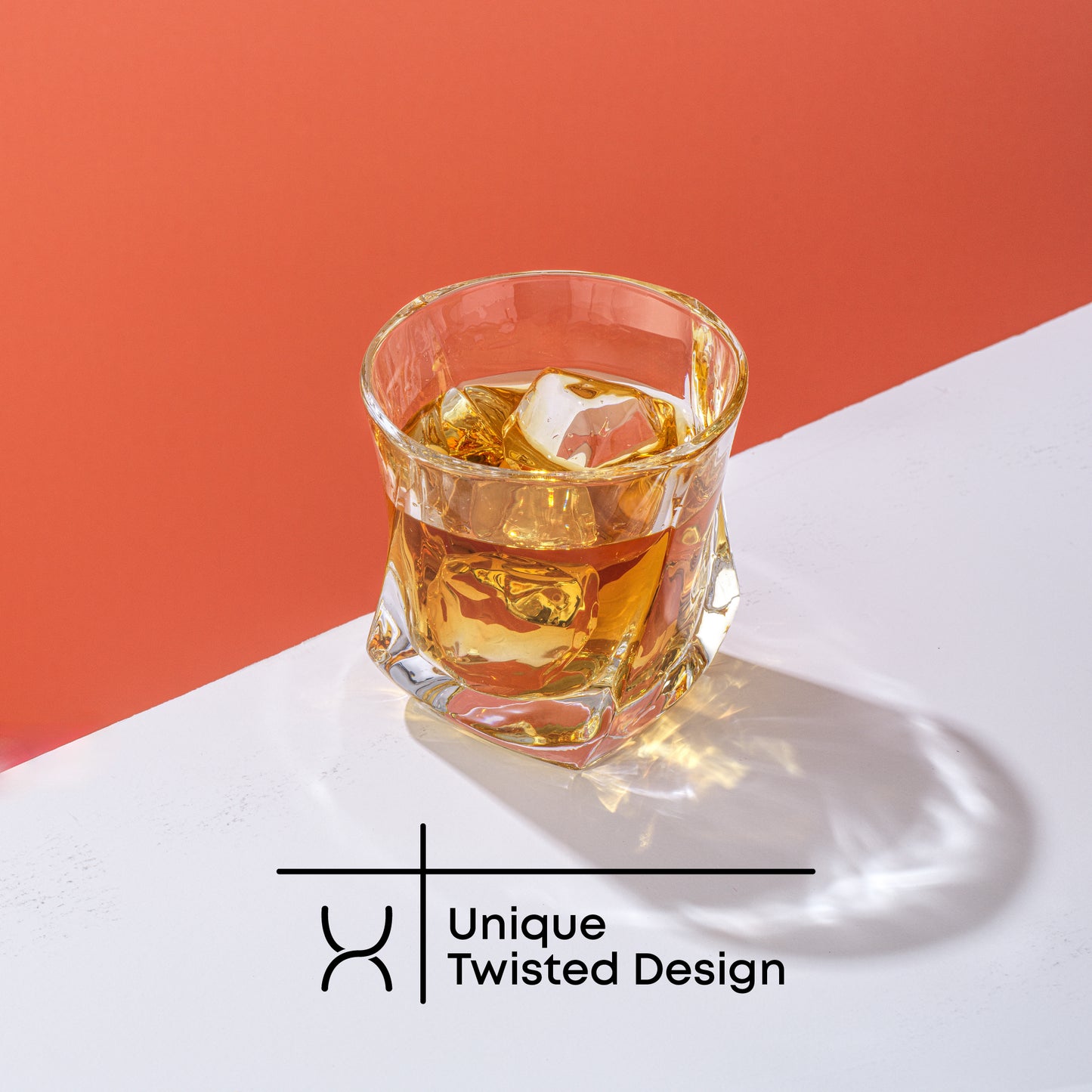 A JoyJolt Aurora Whiskey glass filled with whiskey and ice on a red and white background. Text on image reads, “Unique Twisted Design.”