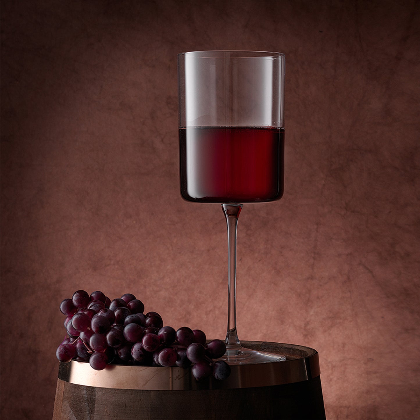 A darker red wine in the wine glass standing on a wooden barrel with grapes next to it