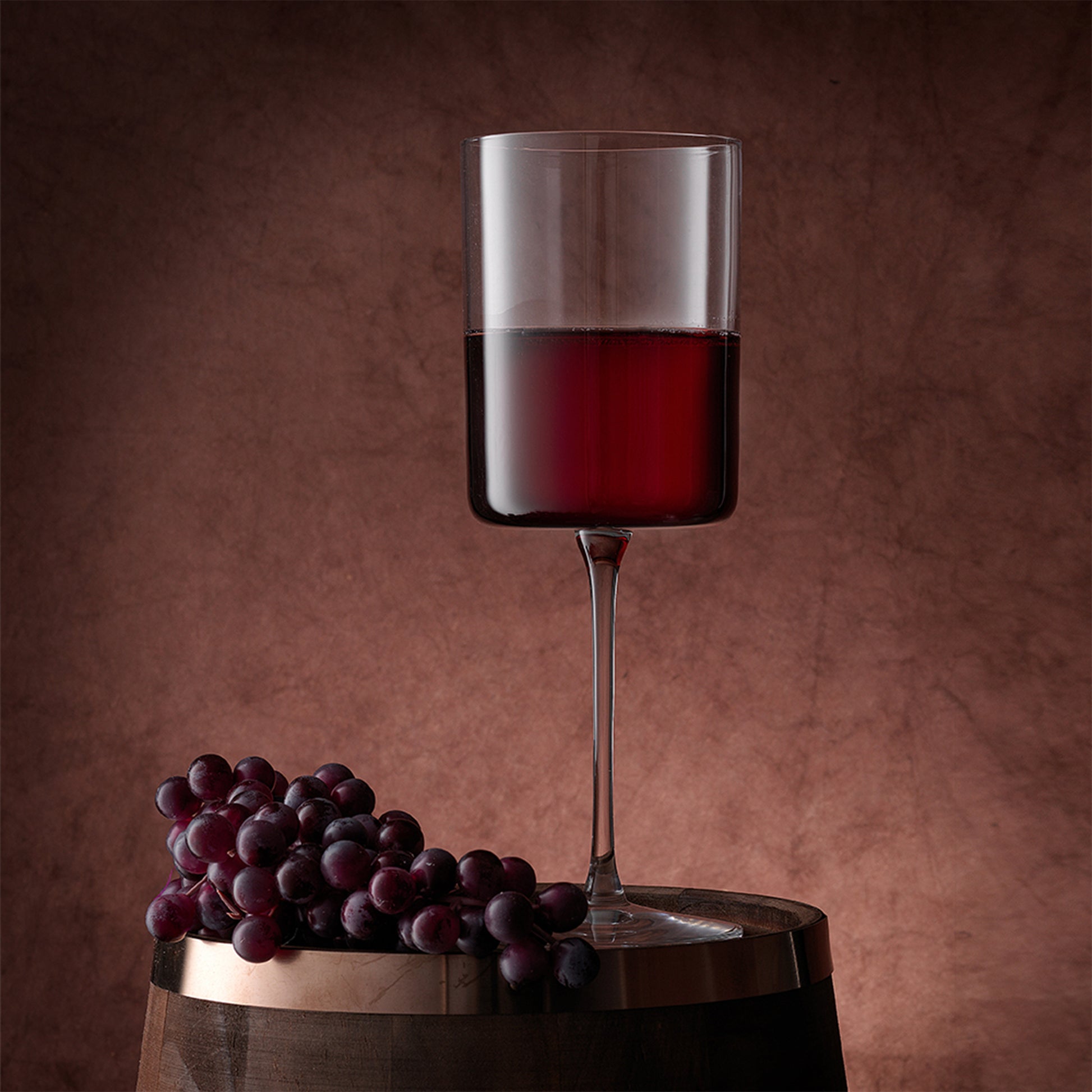 A darker red wine in the wine glass standing on a wooden barrel with grapes next to it