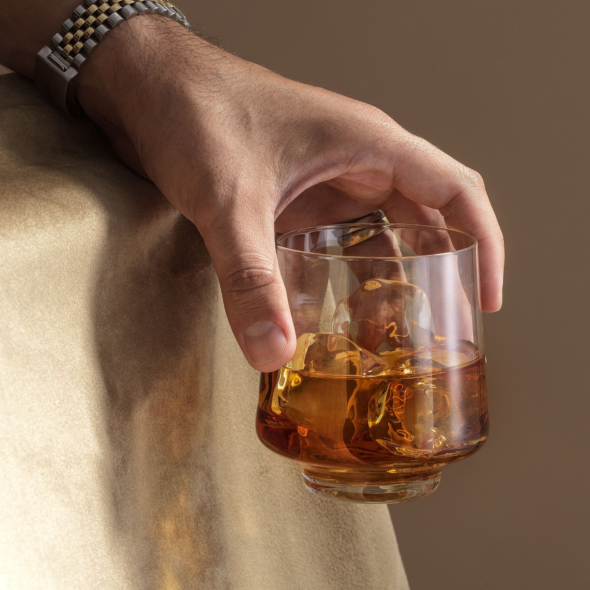 Hand holding a glass of whiskey with ice against a neutral background