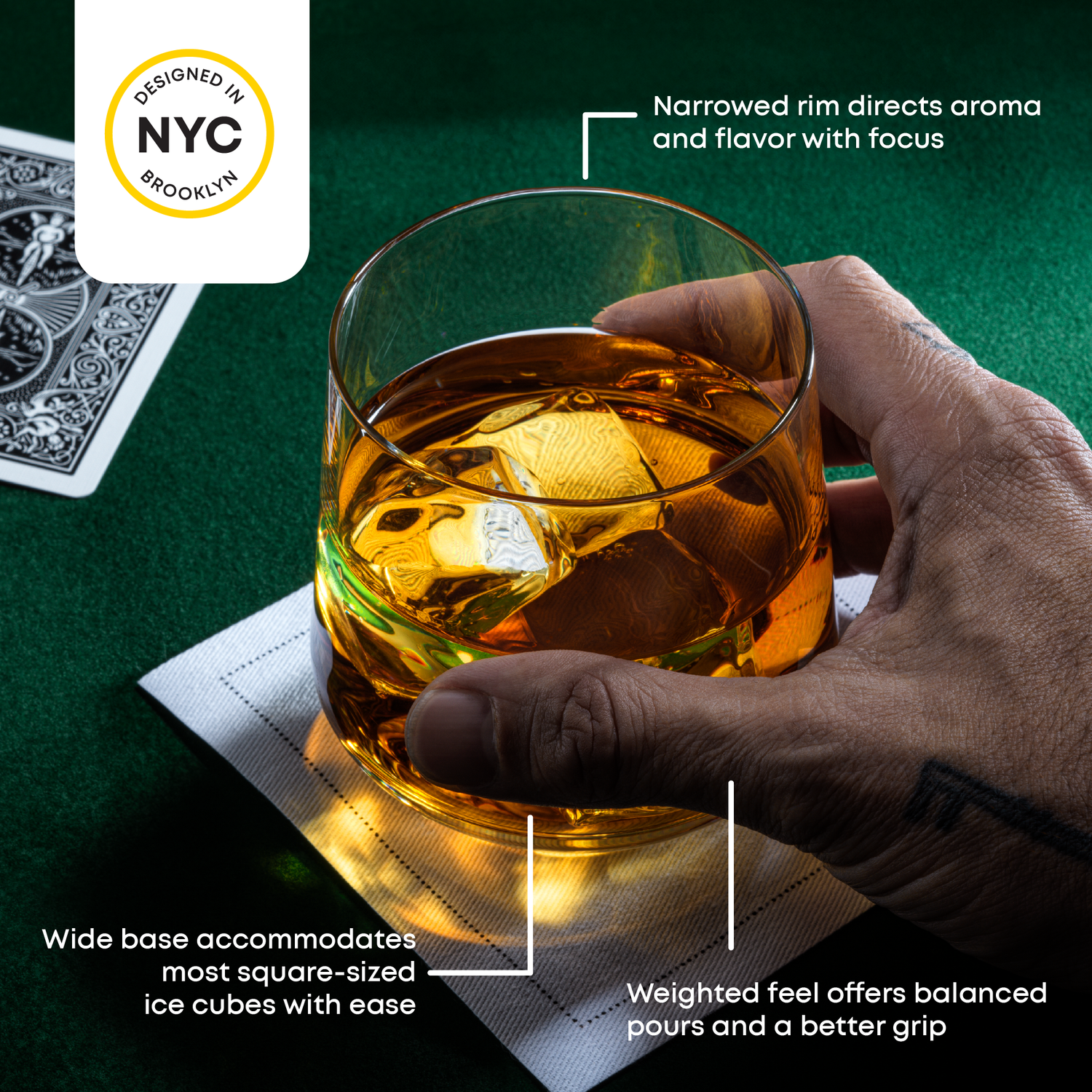 Hand holding a glass of whiskey with ice cubes on a green tablecloth, featuring text about the glass design: "rim directs aroma, shape fits ice cubes, weighted base.
