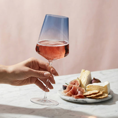 Hand holding a glass of rose wine with a plate of cheese and figs on a marble surface.