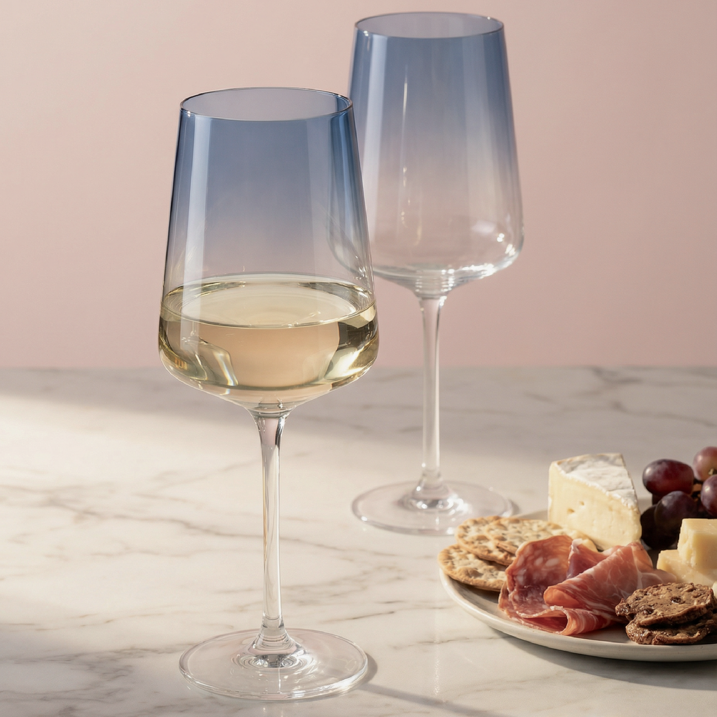 Two wine glasses with white wine on a marble surface with a plate of cheese and snacks.
