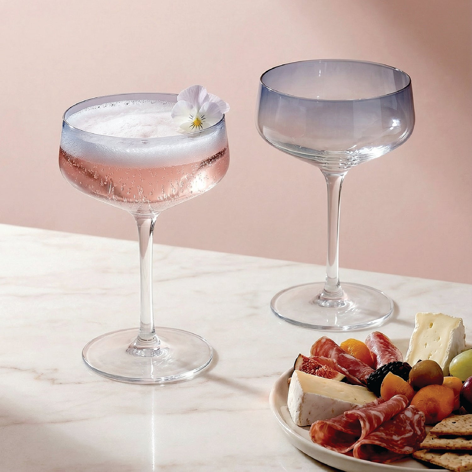 Two cocktail glasses with a pink drink and a white flower, one plain and one with a gradient, on a marble surface with a plate of snacks.