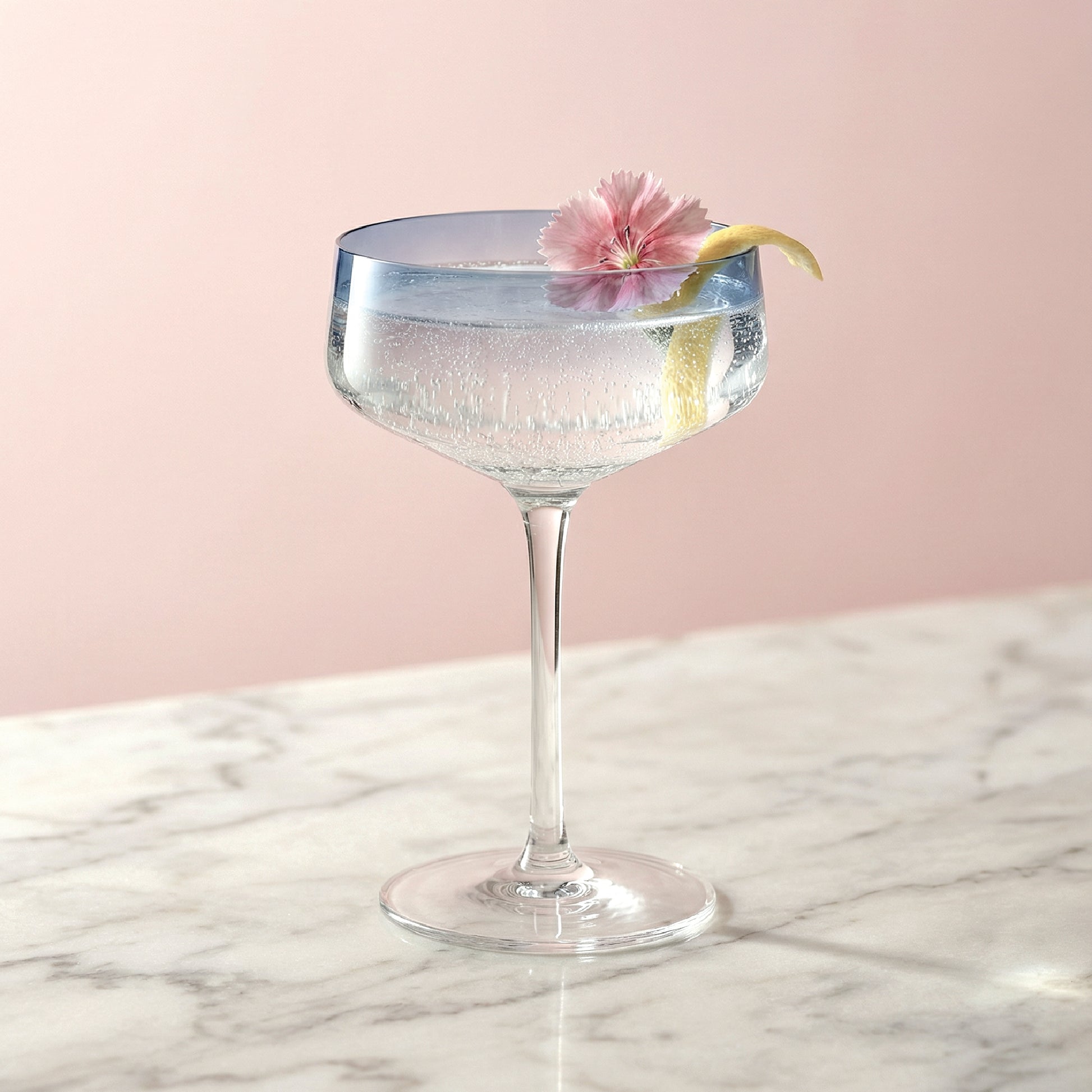 Cocktail in a glass with a pink flower and lemon twist on a marble surface with a pink background