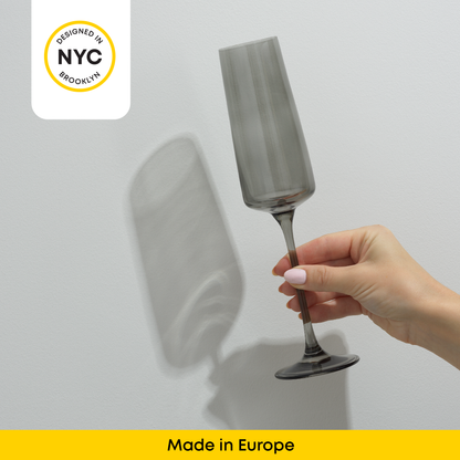 Hand holding a gray champagne glass against a grey background with 'Designed in NYC' and 'Made in Europe' text.