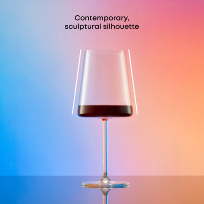 Contemporary Aura red wine glass with sculptural tapered silhouette against a blue, pink, and orange background.