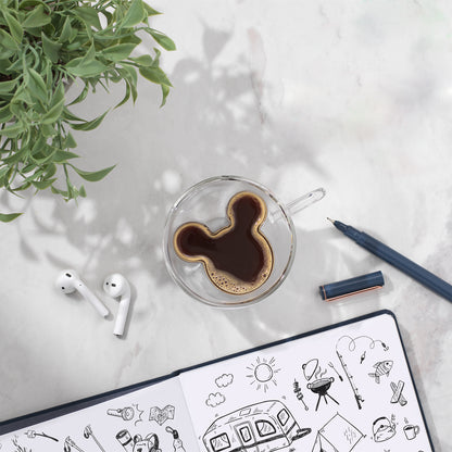 A full Mickey espresso glass next to an notebook with drawings, a plant, and headphones