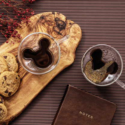 Two Mickey espresso glasses full of espresso with cookies nearby, on a wooden cutting board, with a notebook