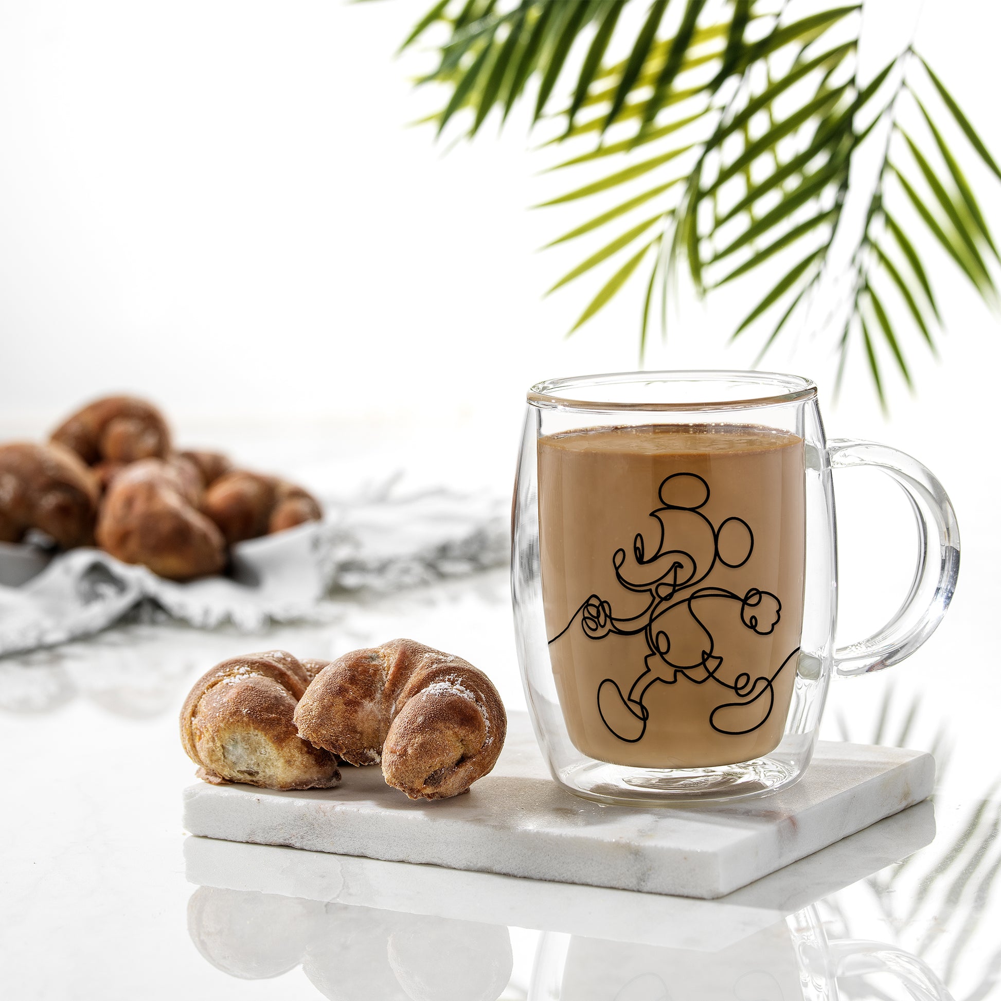 A Mickey mug full of coffee on a coaster with a croissant next to it.