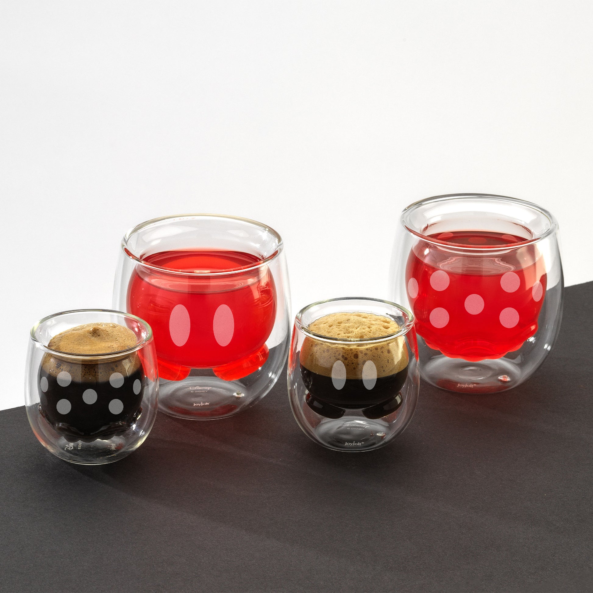 Two sizes of glass cups with a double wall design featuring Mickeys pants and Minnies skirt, filled with a red drink and espresso, on a black surface against a white background