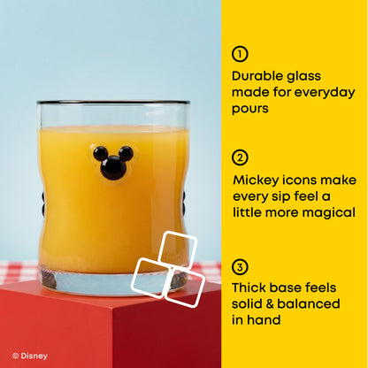 A Mickey Mouse-shaped clear glass tumbler filled with orange juice, shown on a red pedestal against a light blue background with a bright yellow text panel on the right describing product features. Disney branding appears in the lower left.