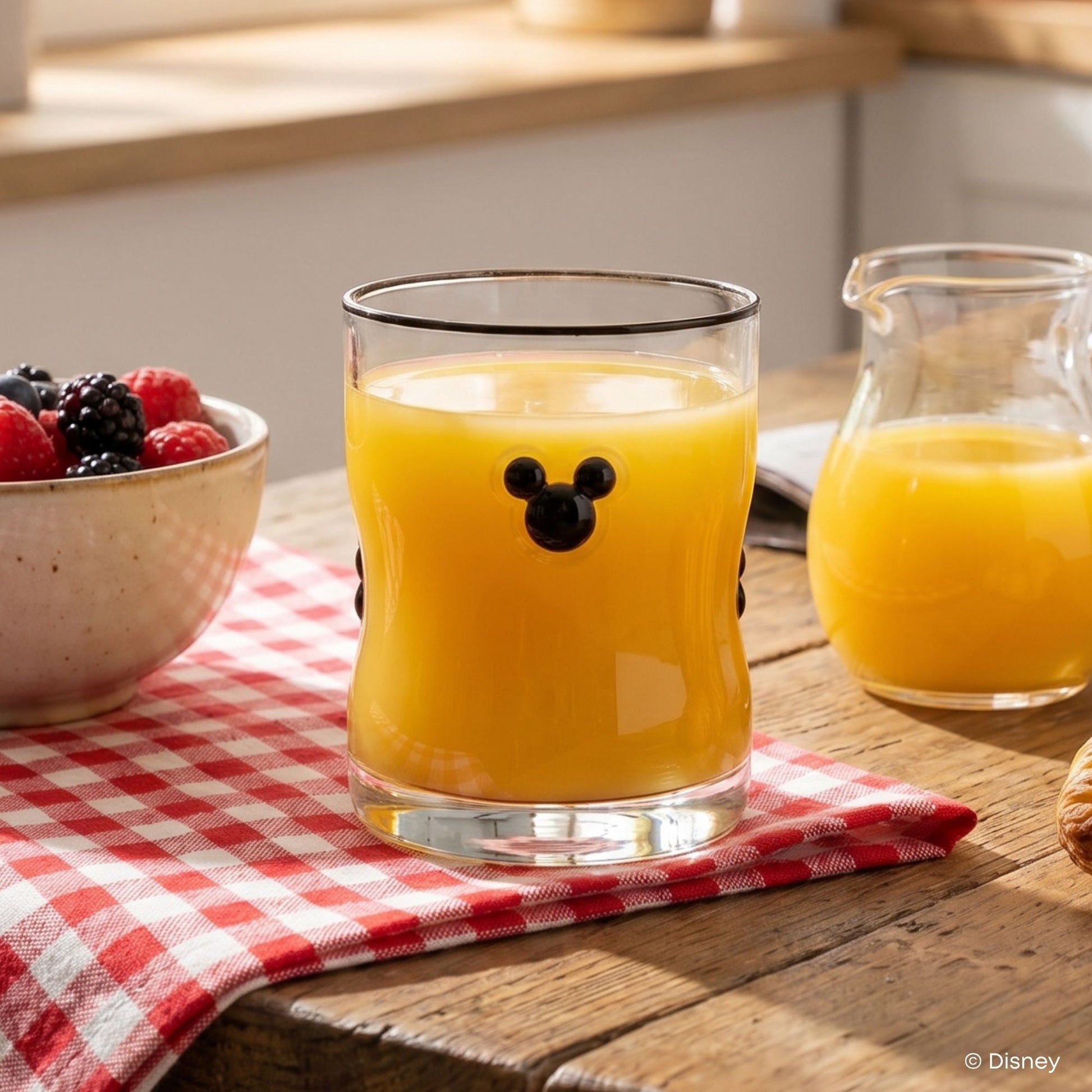 A clear glass tumbler with a black Mickey Mouse head design on the front, filled with orange juice and placed on a red-and-white checkered cloth on a wooden table. A bowl of berries and a small pitcher of orange juice are visible in a kitchen setting.