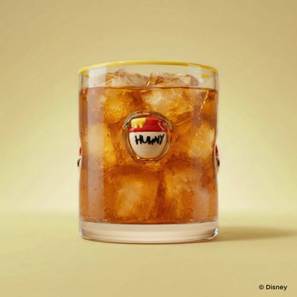 A clear glass tumbler with a yellow rim, filled with iced cola and ice cubes, featuring a circular Winnie the Pooh 'HUNNY' logo on the front. The glass is centered against a warm beige background.