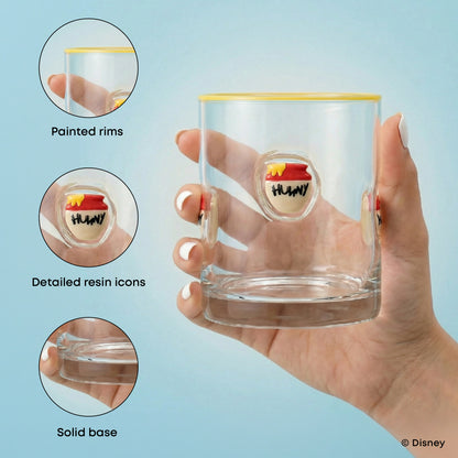 A hand holds a clear drinking glass with a yellow rim and a small multicolor HUNNY logo on the front. Three circular inset callouts on the left highlight product features: painted rims, detailed resin icons, and a solid base.