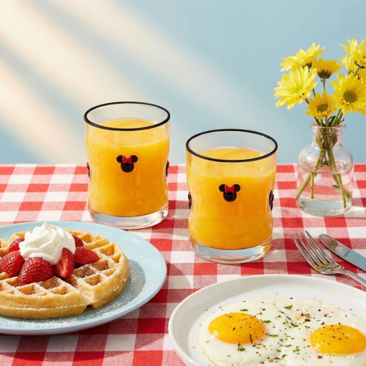 Two clear glass tumblers with black rims and a Minnie Mouse face graphic, filled with orange juice, set on a red-and-white checkered tablecloth with a waffle, strawberries, eggs, and yellow flowers in the background.