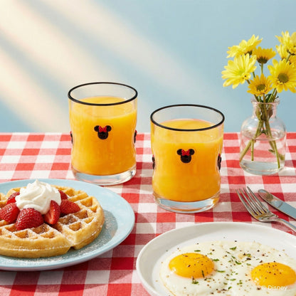 Two clear glass tumblers with black rims and a Minnie Mouse face graphic, filled with orange juice, set on a red-and-white checkered tablecloth with a waffle, strawberries, eggs, and yellow flowers in the background.