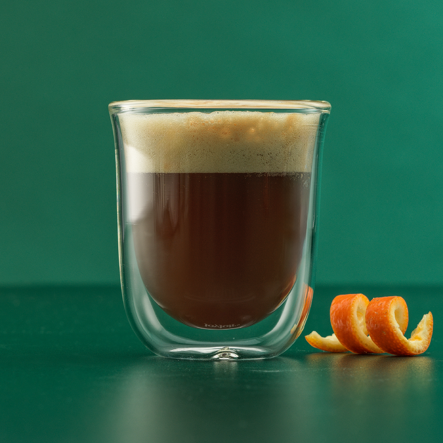 A double wall espresso glass full of espresso with foam against a green surface and green background, with an orange peel next to it.