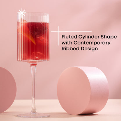 A tall, stemmed fluted glass with filled with a red cocktail. There are two circular shapes next to the glass. The background is pink.