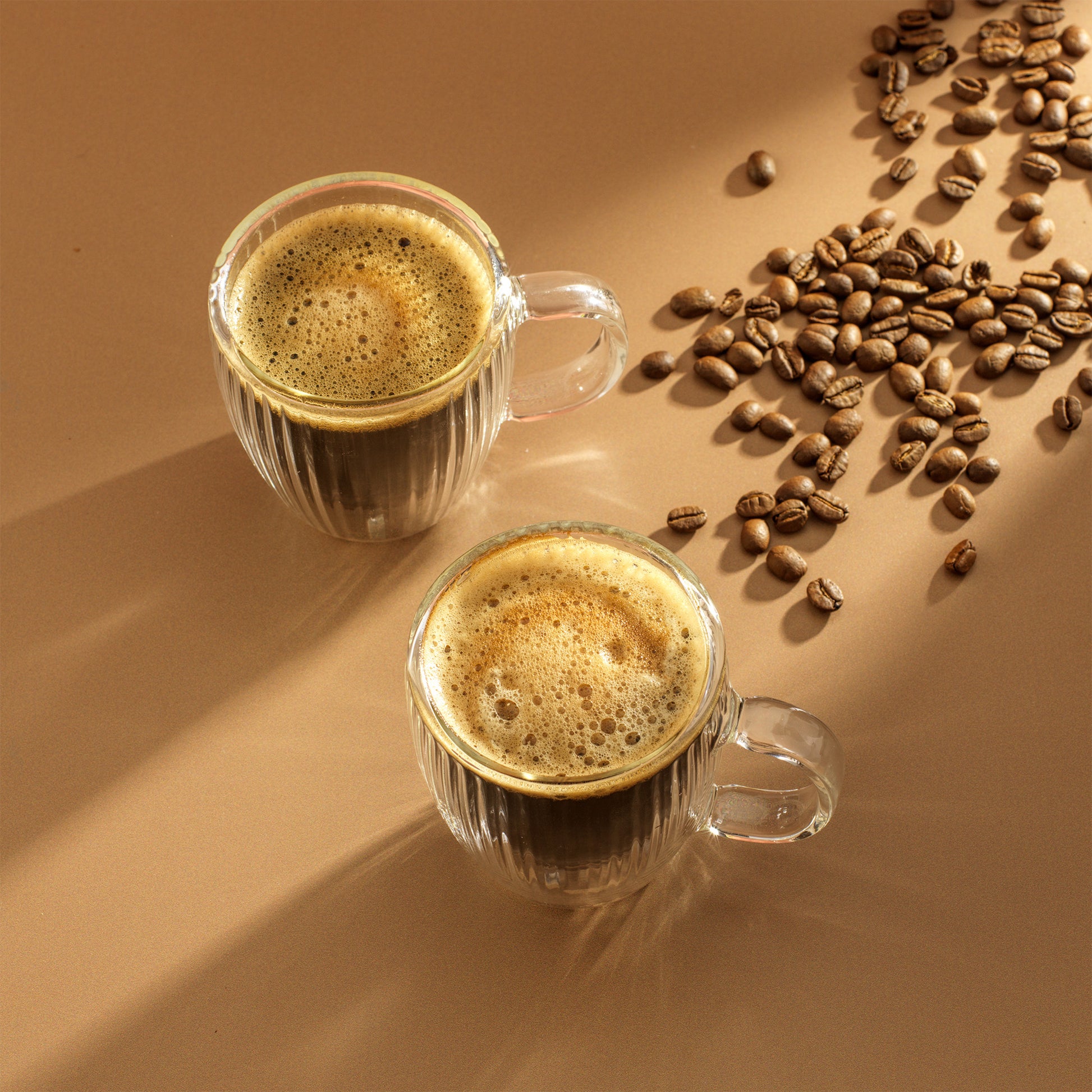 Two fluted glass mugs full of espresso, on a brown surface with coffee beans scattered next to them
