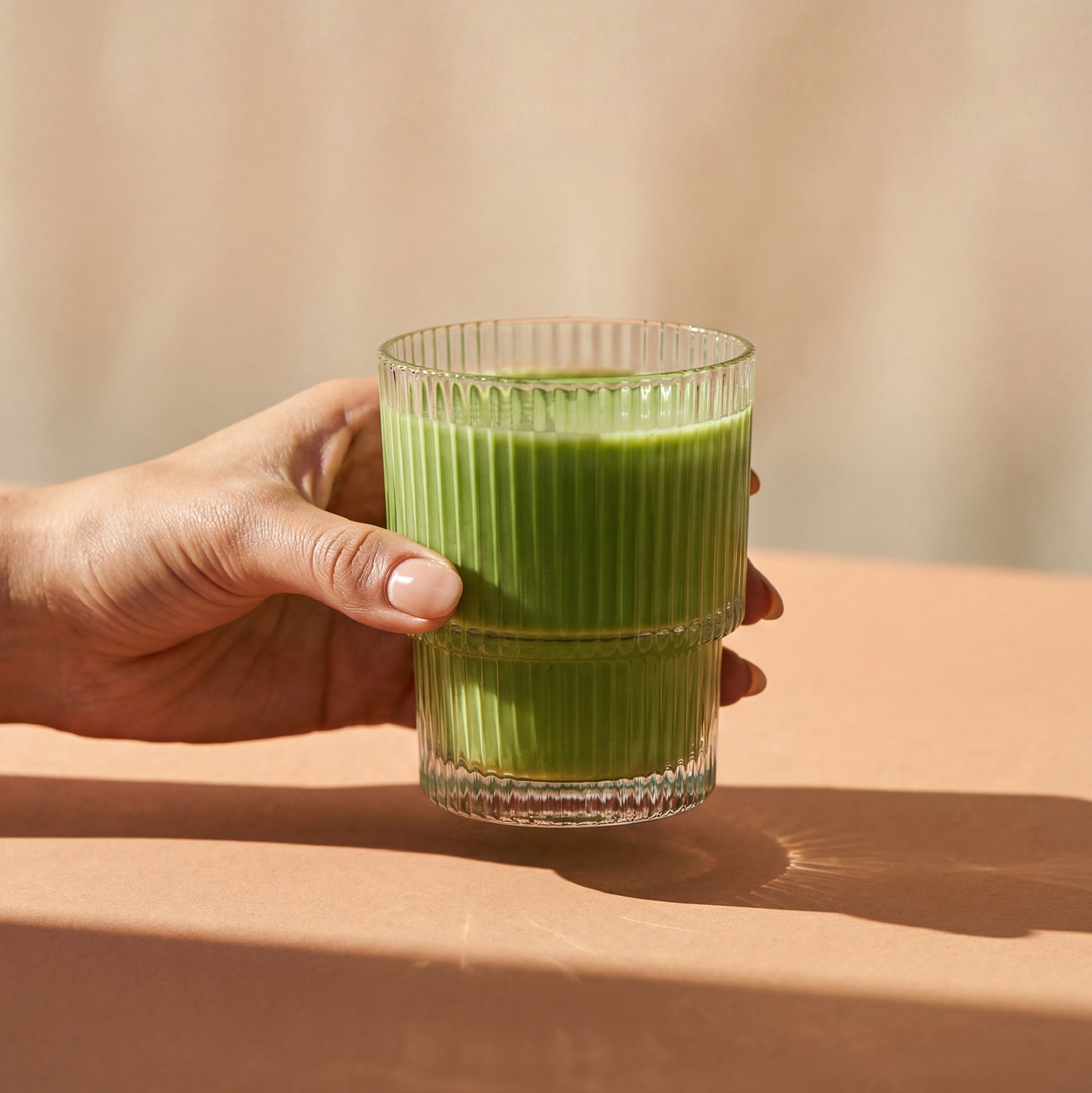 Hand holding a glass of green juice against a neutral background