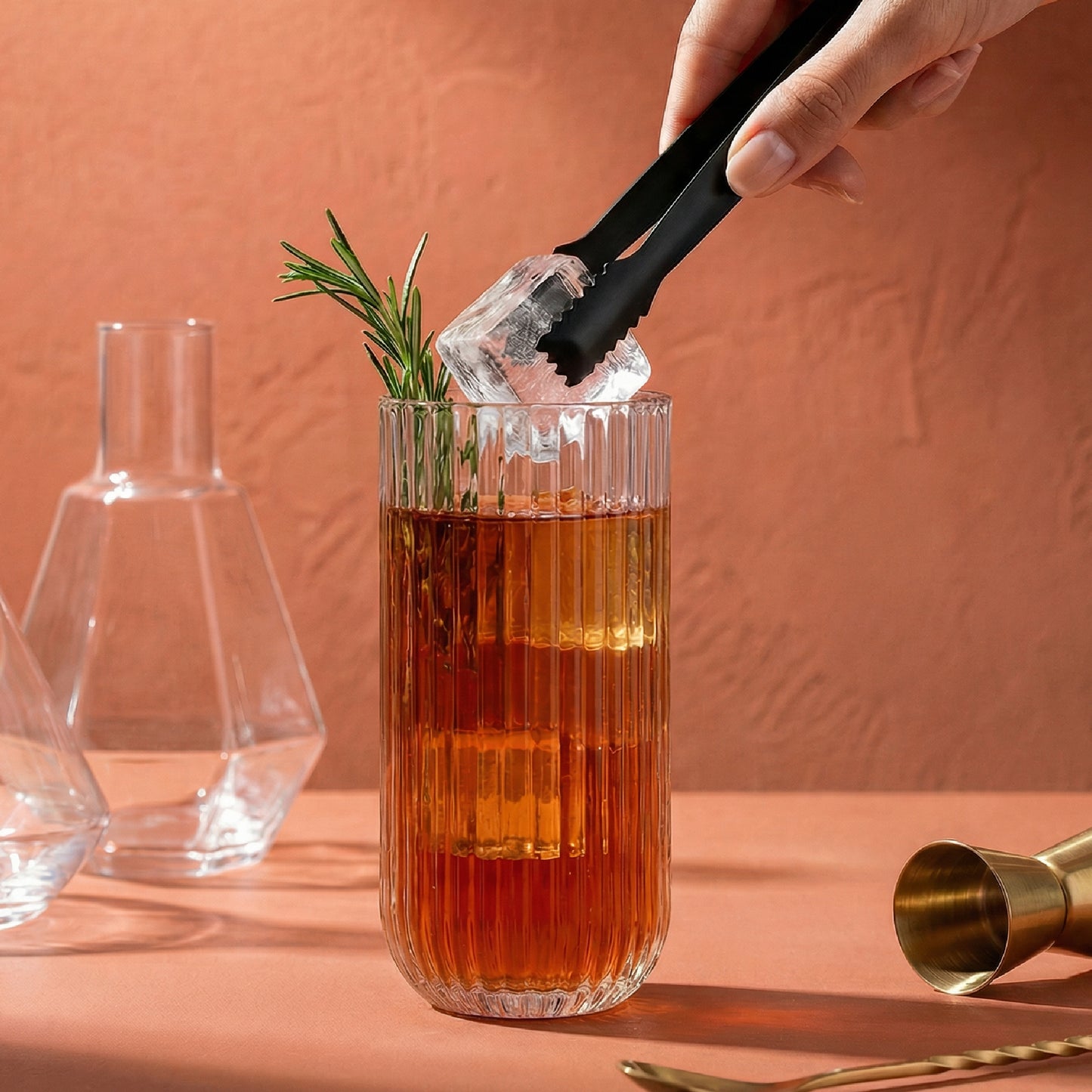 Person adding ice to a cocktail with rosemary garnish in a glass on a pink background
