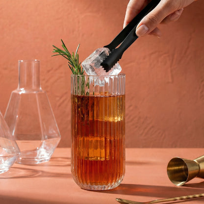 Person adding ice to a cocktail with rosemary garnish in a glass on a pink background