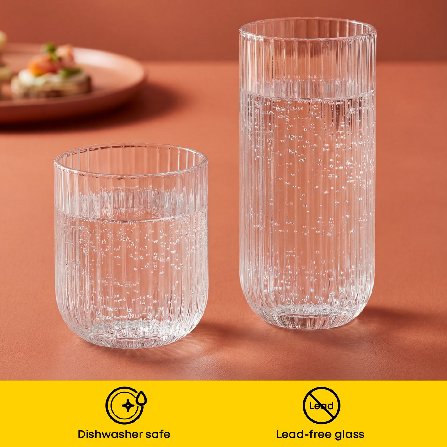 Two clear glass tumblers on a brown surface with a yellow banner at the bottom indicating dishwasher safe and lead-free glass.