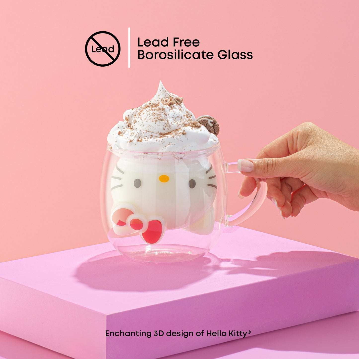 A hand holding a Hello Kitty-themed 3D double wall coffee mug filled with a drink, presented on a pink stand against a pink background. With text "lead free borosilicate glass"