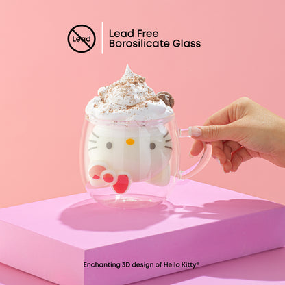 A hand holding a Hello Kitty-themed 3D double wall coffee mug filled with a drink, presented on a pink stand against a pink background. With text "lead free borosilicate glass"