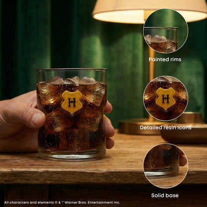 A hand holds a clear lowball-style tumbler glass filled with dark brown iced drink and ice cubes, featuring a yellow shield emblem with a black letter H on the front. The scene is set on a wooden table with a lamp in the background, and three circular inset callouts highlight product details: painted rims, detailed resin icons, and solid base.