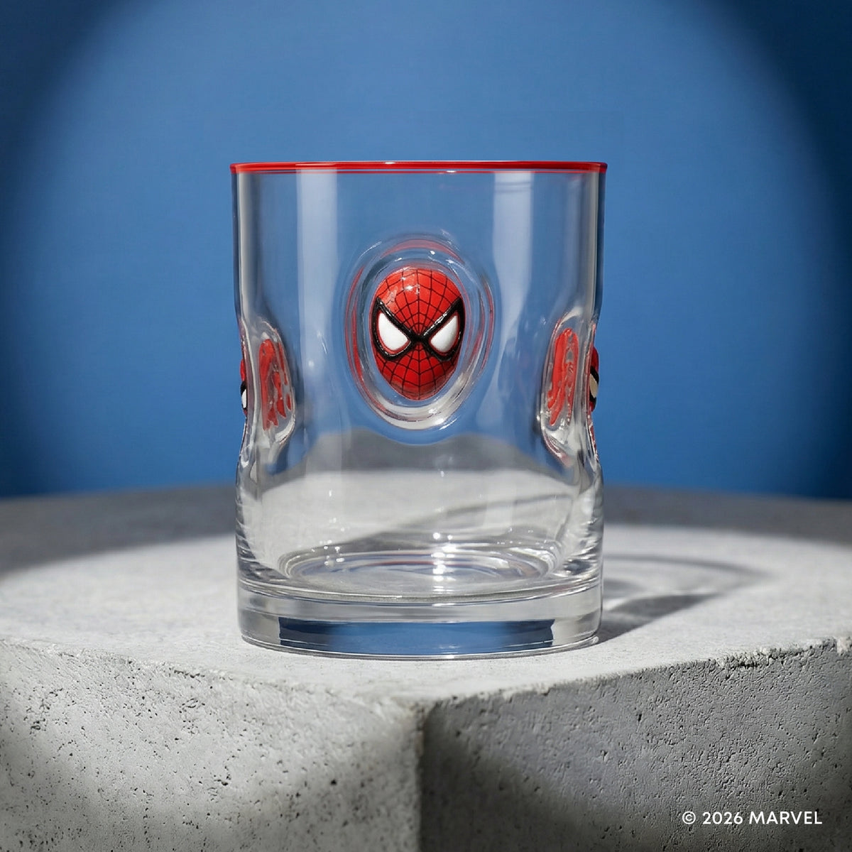 A clear glass tumbler with Spider-Man themed embossed details, featuring a large red and black Spider-Man face in the center and smaller matching faces on the sides, with a red rim, set on a gray concrete pedestal against a blue background.