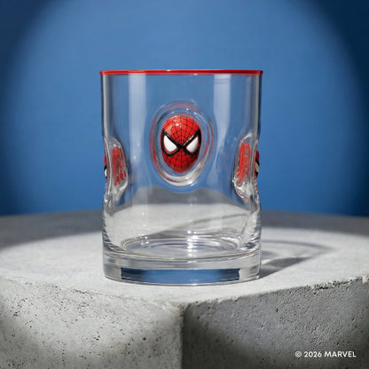 A clear glass tumbler with Spider-Man themed embossed details, featuring a large red and black Spider-Man face in the center and smaller matching faces on the sides, with a red rim, set on a gray concrete pedestal against a blue background.