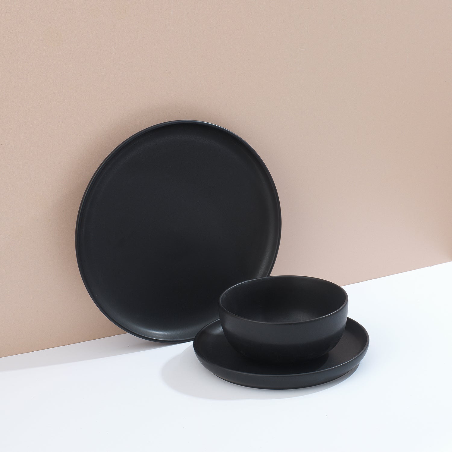 Set of black dinnerware including bowls and plates on a white surface and muted pink background.