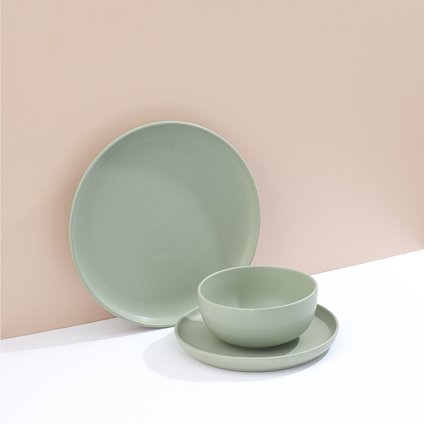 Set of green dinnerware including bowls and plates on a white surface and muted pink background.