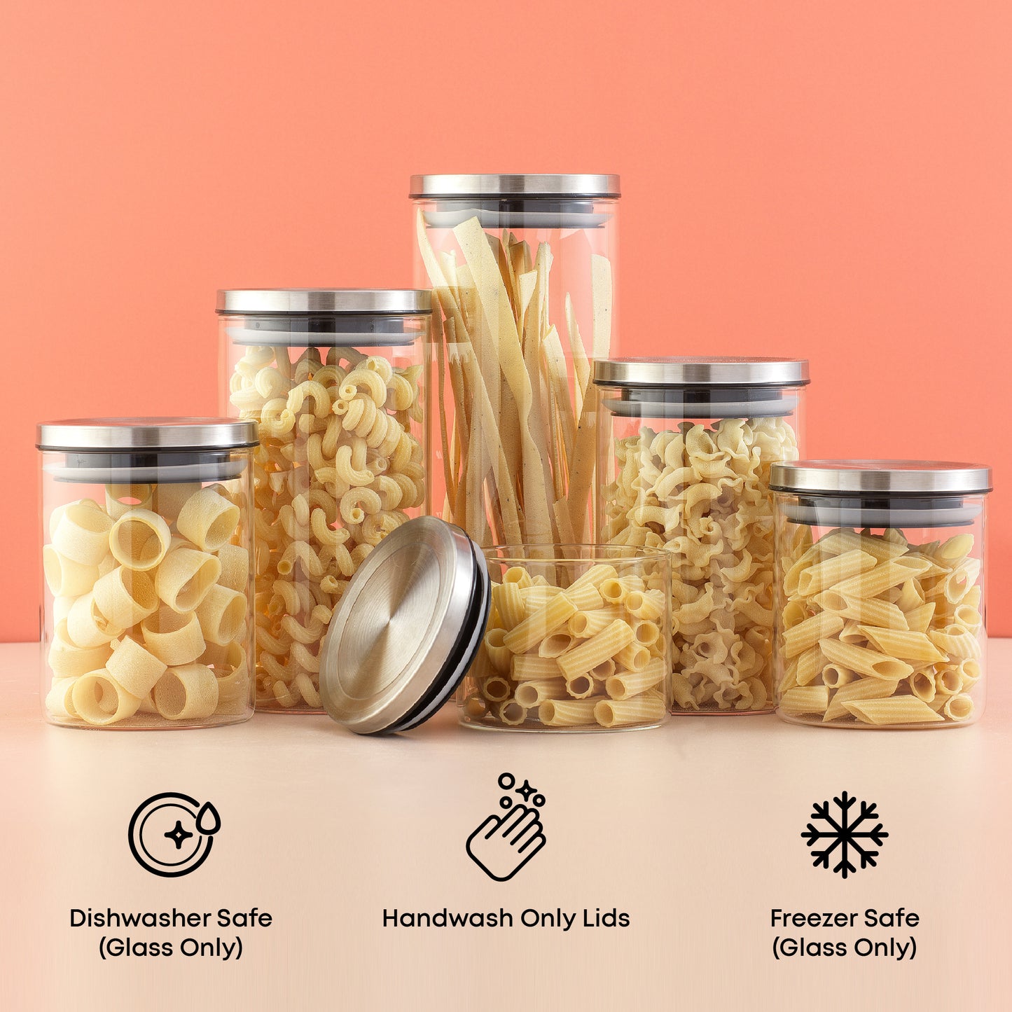 Set of food storage containers with different types of dried pasta on a pink surface with a light red background. Text call outs: dishwasher safe glass, handwash lids, freezer safe glass.
