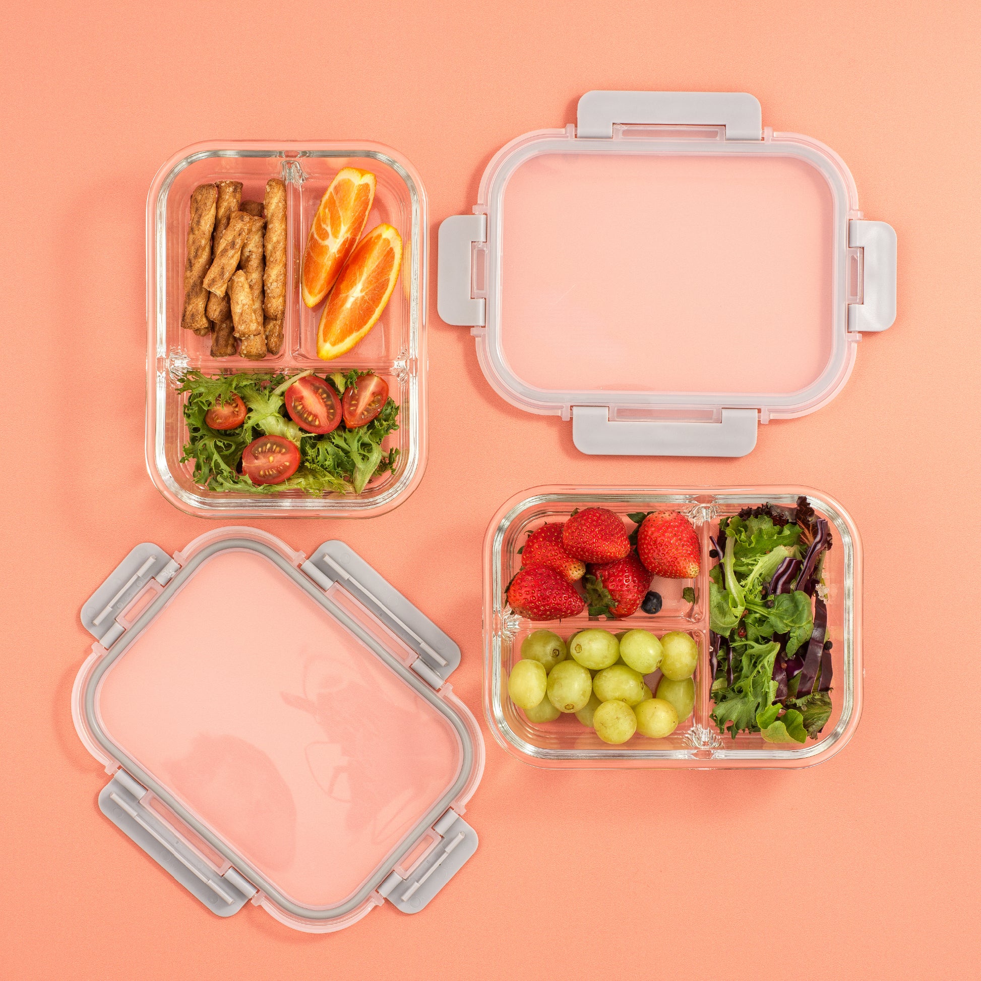 Clear food storage containers with compartments filled with fruits, vegetables, and snacks on a pink background
