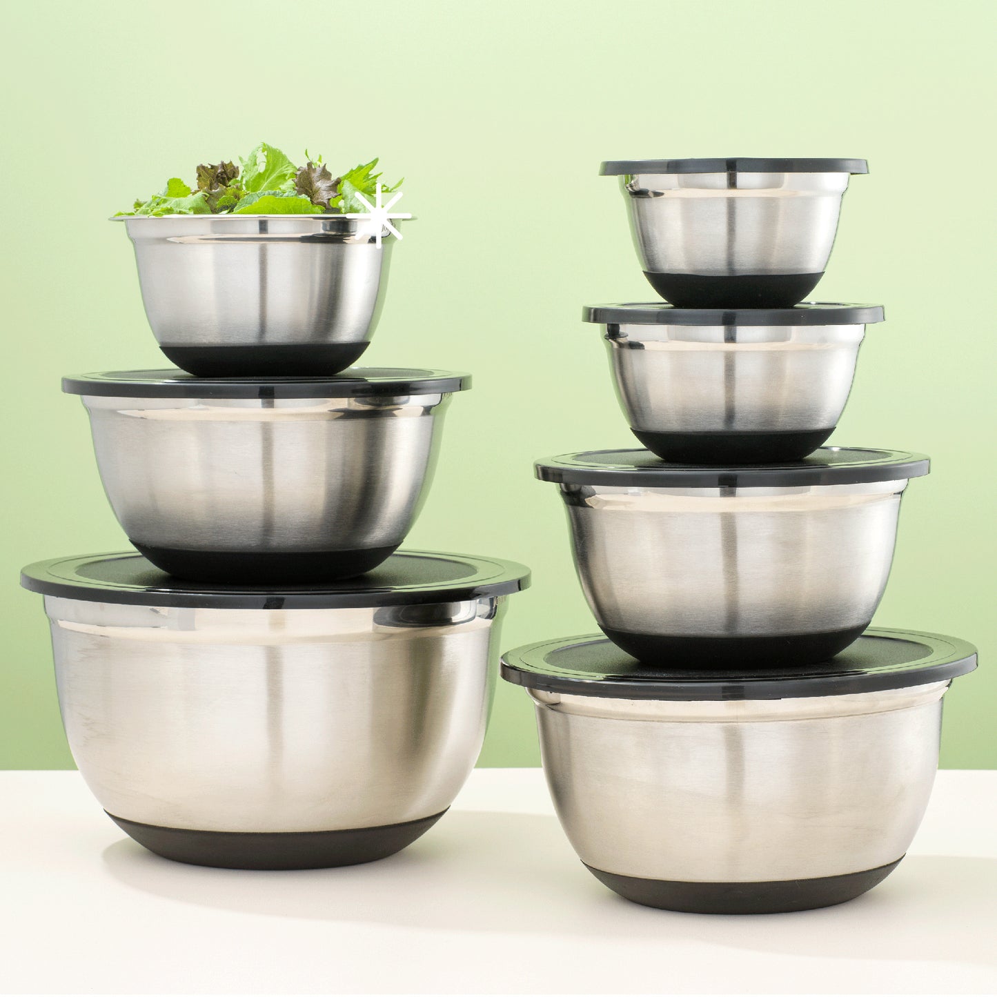 Set of stainless steel mixing bowls with black lids on a white surface with green background. Medium-small size bowl contains a salad.