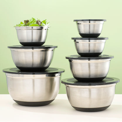 Set of stainless steel mixing bowls with black lids on a white surface with green background. Medium-small size bowl contains a salad.