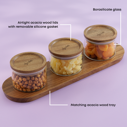 Three glass canisters with wooden lids on a matching wooden tray against a light purple background. with text "borosilicate glass, airtight acacia wood lids, matching acacia wood tray"