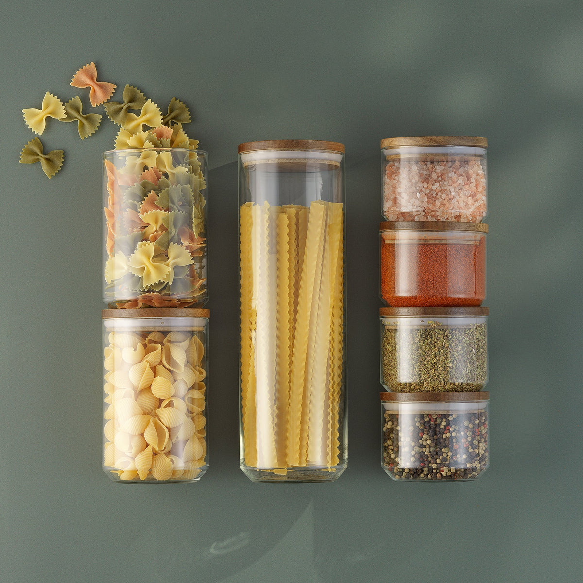 Holden glass food storage jars with wooden lids arranged with pasta, pink salt, spices, and herbs against a muted green background.