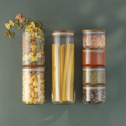 Holden glass food storage jars with wooden lids arranged with pasta, pink salt, spices, and herbs against a muted green background.